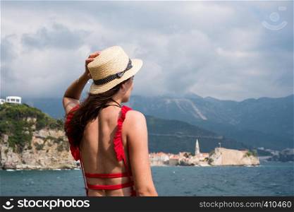 Back view of a young beautiful woman in a hat looking to the old town Budva, Montenegro. Summer vacation destination. Back view of a young beautiful woman in a hat looking to the old town Budva, Montenegro