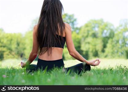 Back view of a young woman doing yoga meditation in a lotus pose outdoors in a park on a sunny day