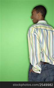 Back view of African-American smiling teen boy with hand in back pocket of jeans standing against green background.