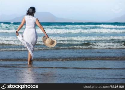 Back view of anonymous woman in white dress carrying straw hat and walking in waves of stormy sea against blue sky while spending summer vacation on beach. Female traveler walking near sea waves