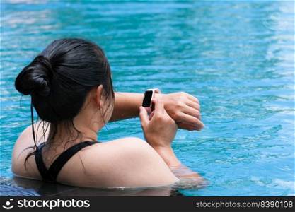 Back view of Asian woman in swimsuit looking at smartwatch after taking a break from swimming in the swimming pool for exercise.