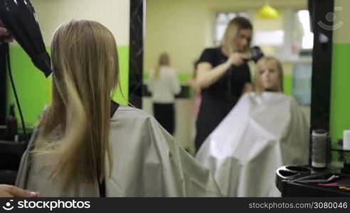 Back view of attractive hairstylist drying cute girl&acute;s hair with hair dryer in barber shop. Rack focus. Positive beautician giving a new hairstyle to child in beauty salon.