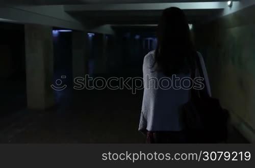 Back view of attractive woman, scared by a stranger, running through dark underground passage . Frightened young girl running away from stranger through underpass at night. Slow motion. Steadicam stabilized shot.