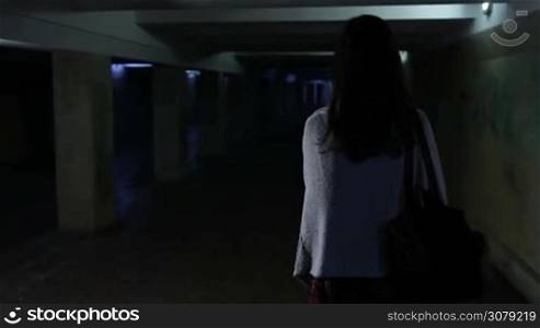 Back view of attractive woman, scared by a stranger, running through dark underground passage . Frightened young girl running away from stranger through underpass at night. Slow motion. Steadicam stabilized shot.
