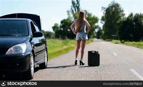Back view of beautiful girl on high heels and shorts standing on a rural road with canister near her broken car and hitchhiking