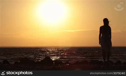 Back view of beautiful slender girl&acute;s silhouette in summer dress enjoying amazing orange sunset as she stands on seashore on summer windy day. Slow motion. Charming serene long blonde hair woman in pure happiness and elated enjoyment watching sundown