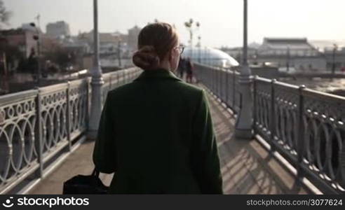 Back view of beautiful woman walking on iron bridge over cityscape background. Rear view of businesswoman strolling alone pedestrian bridge in city. Slow motion.