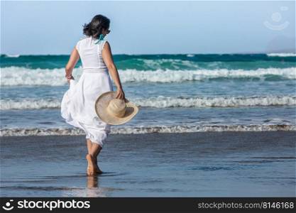Back view of brunette in white dress with straw hat walking on wet beach towards foamy sea waves on summer day on resort. Female tourist walking towards waving sea