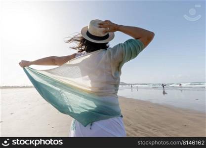 Back view of brunette with thin scarf touching straw hat and enjoying freedom while spending windy weekend day on sandy beach near sea. Unrecognizable female enjoying freedom on beach