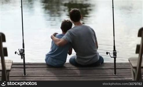 Back view of caring father embracing his son while sitting on wooden pier by the lake angling in summer. Affectionate dad hugging teenage boy and chatting while fishing together with rods at freshwater pond over calm water surface background.