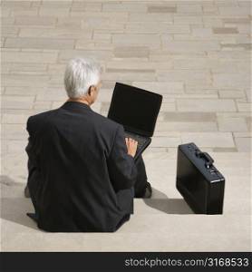 Back view of Caucasian middle aged businessman sitting on steps outdoors with laptop and briefcase.