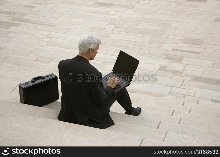 Back view of Caucasian middle aged businessman typing on laptop with briefcase sitting on steps outdoors.