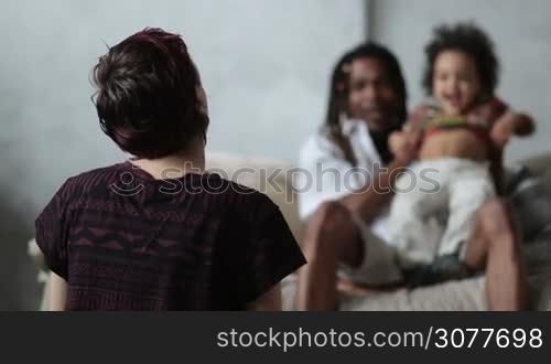 Back view of caucasian mother sitting on the floor while african american dad with dreadlocks playing with their little mixed race toddler son. Cheerful diverse family spending great time together at home. Selective focus. Slow motion.