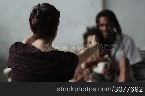 Back view of caucasian mother sitting on the floor with open arms, waiting for mixed race cute child to embrace. Blurry african american father with dreadlocks sitting on sofa and holding little boy. Cute kid runs away from mom&acute;s embrace. Slow motion