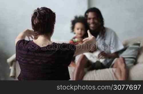 Back view of caucasian mother with open arms sitting on the floor to embrace her cute mixed race toddler son over blurry african american father sitting on the sofa with little child background. Slow motion.