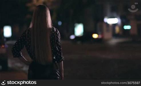 Back view of charming girl hitchhiking on the city street at night. Background big city lights