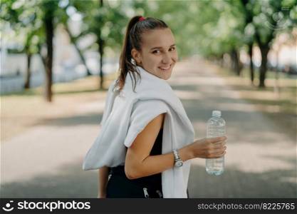 Back view of cheerful glad sporty woman with pony tail stays hydrated after cardio workout in park holds bottle of fresh water takes break after exhausting morning jogging goes in for sport regularly