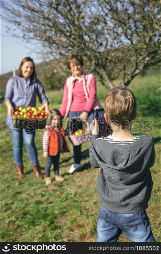 Back view of cute boy taking a photo with electronic tablet to family with fresh organic apples in a wicker basket after harvest. Family leisure time concept.. Boy taking photo to family with apples in basket