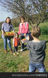 Back view of cute boy taking a photo with electronic tablet to family with fresh organic apples in a wicker basket after harvest. Family leisure time concept.. Boy taking photo to family with apples in basket