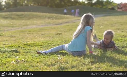 Back view of cute teenage girl sitting on green grass and playing with her laughing toddler brother. Smiling little boy having fun and laughing while her sister tickling him. Excited siblings enjoying leisure in park in glow of beautiful sunset.