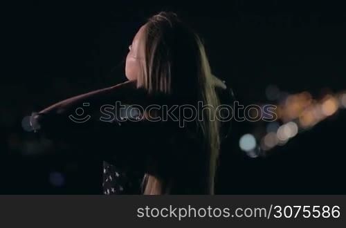 Back view of cute woman is standing outdoors on a cool summer &acute;s night and wind is playing with her blonde long hair. Shallow depth of field. Focus on woman. Background cityscape and streetlights at night