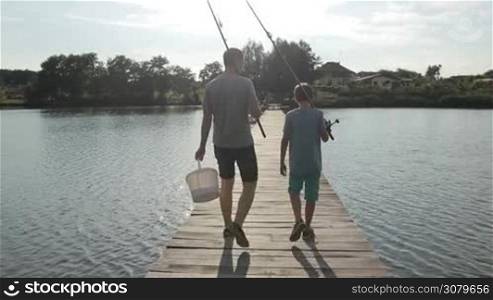 Back view of dad carrying bucket in one hand and teenage boy with fishing rod walking along wooden pontoon for fishing in rays of setting sun. Relaxed father and son with spinning rods going fishing on pond over awesome rural landscape background.