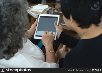 Back view of elderly old senior asian woman using health monitoring application on digital tablet while gathered together with grandson in living room.
