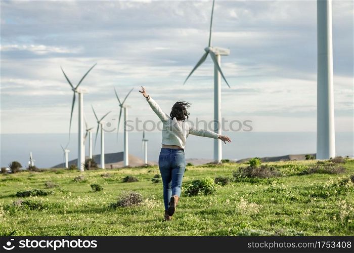 Back view of faceless carefree female running with outstretched arms in field with windmills and enjoying freedom on sunny day on Lanzarote. Unrecognizable woman running in meadow with windmills