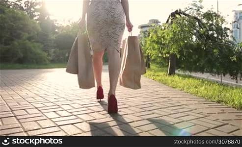 Back view of fashion shopper woman&acute;s legs in red high heels with shopping bags walking in the street. Close-up. Low section of elegant female carrying shopping bags while stepping along the street at sunset. Slow motion. Steadicam stabilized shot.