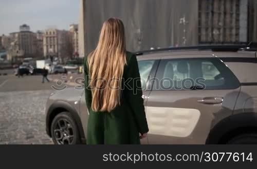Back view of fashionable female driver with long blonde hair wearing emerald green coat opening car&acute;s door and getting in the automobile. Rear view of attractive woman getting into parked car in daylight over urbanscape background. Slow motion.