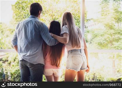 back view of father and mother with daughter standing at window and looking outside at home, happy young family concept