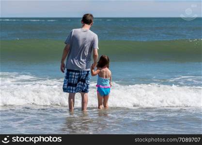 Back view of father and young girl facing the sea waves as concept for protection against future problems. Rear view of father and young daughter facing the ocean waves