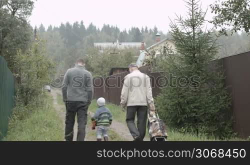 Back view of father, son and grandpa walking along the road in the village. Old man rolling a trolley bag.