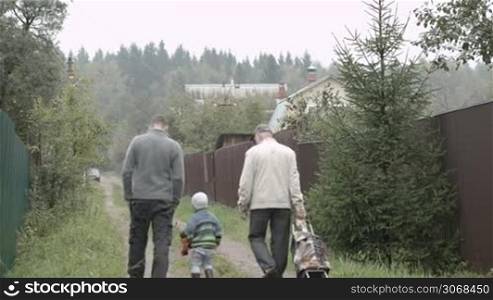 Back view of father, son and grandpa walking along the road in the village. Old man rolling a trolley bag.