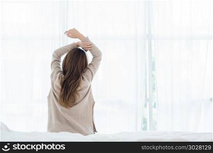 Back view of happy beautiful young Asian woman waking up in morn. Back view of happy beautiful young Asian woman waking up in morning, sitting on bed, stretching in cozy bedroom, looking through window. Funny woman after wake up. She is stretching and smiling.