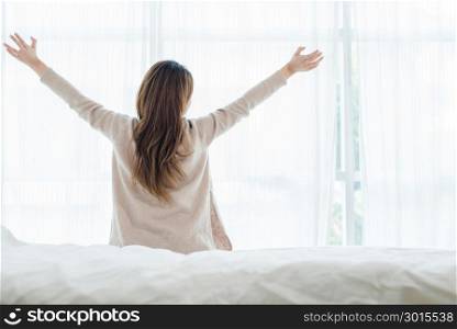 Back view of happy beautiful young Asian woman waking up in morning, sitting on bed, stretching in cozy bedroom, looking through window. Funny woman after wake up. She is stretching and smiling.