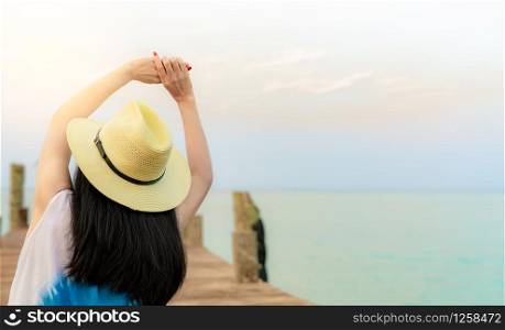 Back view of happy young Asian woman in casual style fashion and straw hat relax and enjoy holiday at tropical paradise beach. Girl sit at the wood pier of resort in summer vacation. Summer vibes.