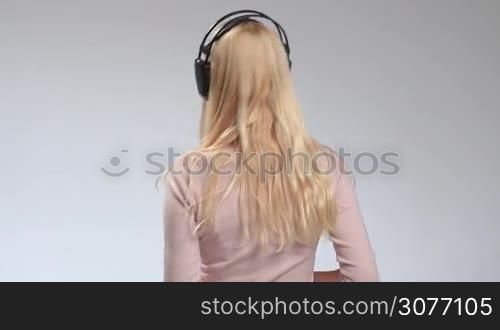 Back view of long hair blonde girl wearing earphones and swaying to the beat on white. Young stunning woman turning back and looking at the camera with charming smile while enjoying the music streaming on big headphones.