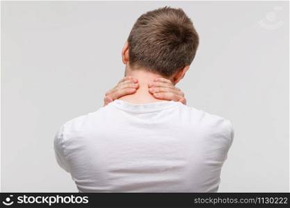 Back view of man in white top touching her pain in his neck and back, feels pain, massages the painful area. Cervical arthritis, osteochondrosis, diseases of musculoskeletal system