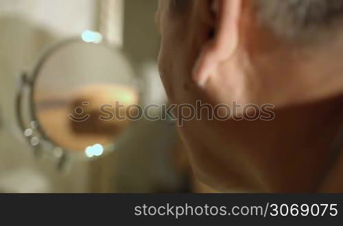 Back view of mature man shaving in the bathroom with his reflection in small round mirror