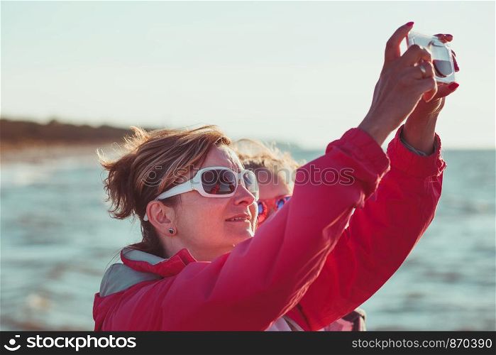 Back view of mother and her daughter taking a photos over sea during summer vacation using smartphone camera