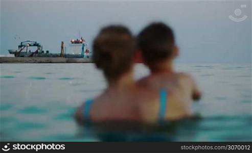 Back view of mother and son in sea looking at dock with ships in the evening