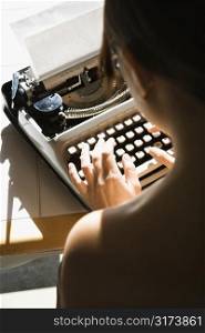 Back view of nude young Asian woman sitting at kitchen table typing on typewriter.
