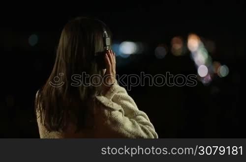 Back view of passionate young female in headphones listening to music while enjoying streetlights at night. Joyful woman holding hands on earcups, swinging along to beat of music while relaxing in park with blurred city lights on background.