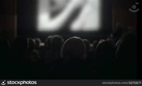 Back view of people watching movie in the cinema. Defocused screen with film lightening viewers in dark hall