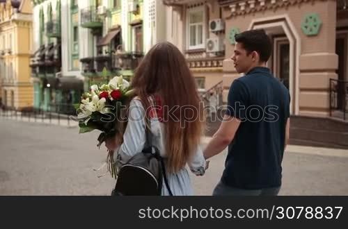 Back view of positive couple in love spending time together while walking down city street on romantic date. Angle view. Young dating couple holding hands strolling along street, chatting and enjoying leisure on sunny day. Slow motion. Stabilized shot.