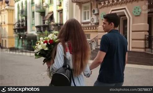 Back view of positive couple in love spending time together while walking down city street on romantic date. Angle view. Young dating couple holding hands strolling along street, chatting and enjoying leisure on sunny day. Slow motion. Stabilized shot.