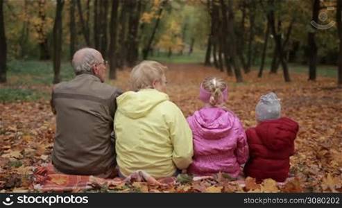 Back view of positive multi-generation family embracing each other while sitting on yellow fallen leaves in the park over colorful autumn background. Grandparents and grandchildren spending time together on fresh air as they walk in park in fall.