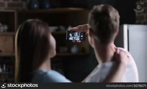 Back view of positive young couple standing embraced and making self-portrait with mobile phone in domestic kitchen. Focus on image displaying on the smartphone screen.
