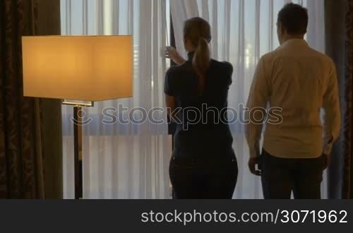 Back view of romantic couple at home or in hotel room. Woman opening the curtain, they embracing gently and looking out the window. Happy time together